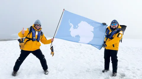 Two people in yellow jackets holding up an Antarctica flag in the snow.